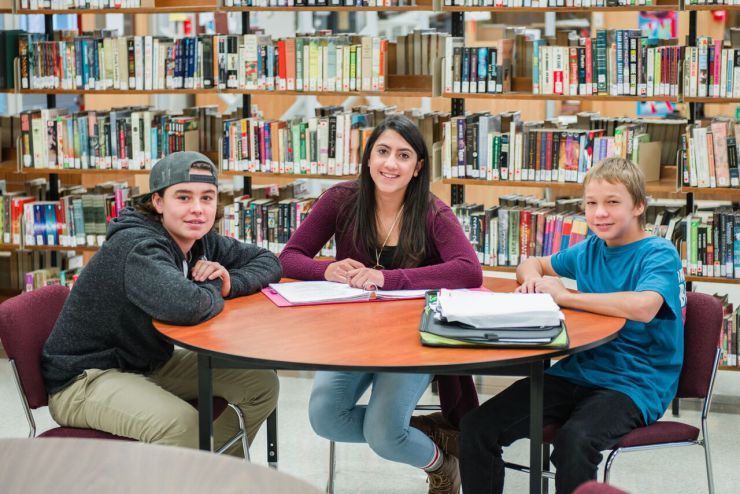 Three students sit at a table in a library with notebooks