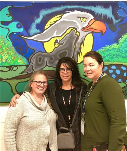 Three women pose in front of an Indigenous art mural 