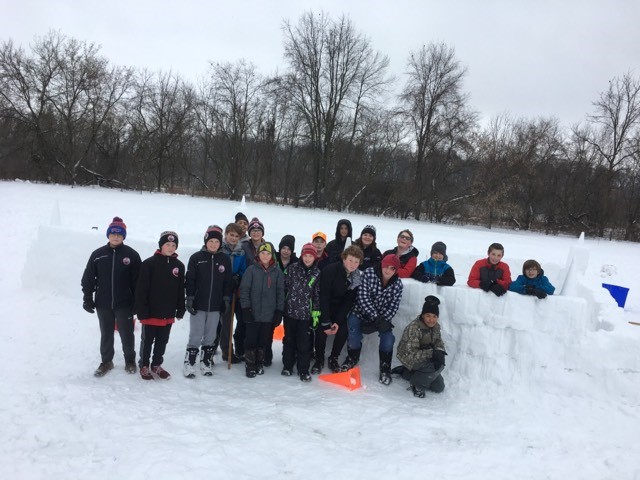 A group of students wearing snowsuits stands in front of a snow fort