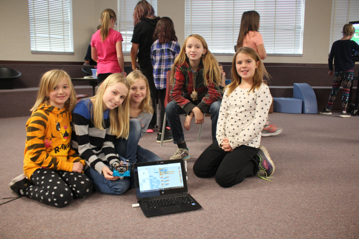 A small group of girls poses with a laptop and robot