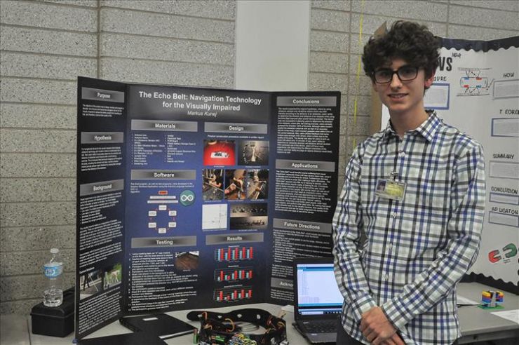 A male student with glasses smiles while standing in front of science fair display board