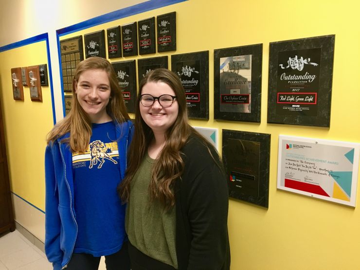 Two female students pose in front of a wall of theatre awards